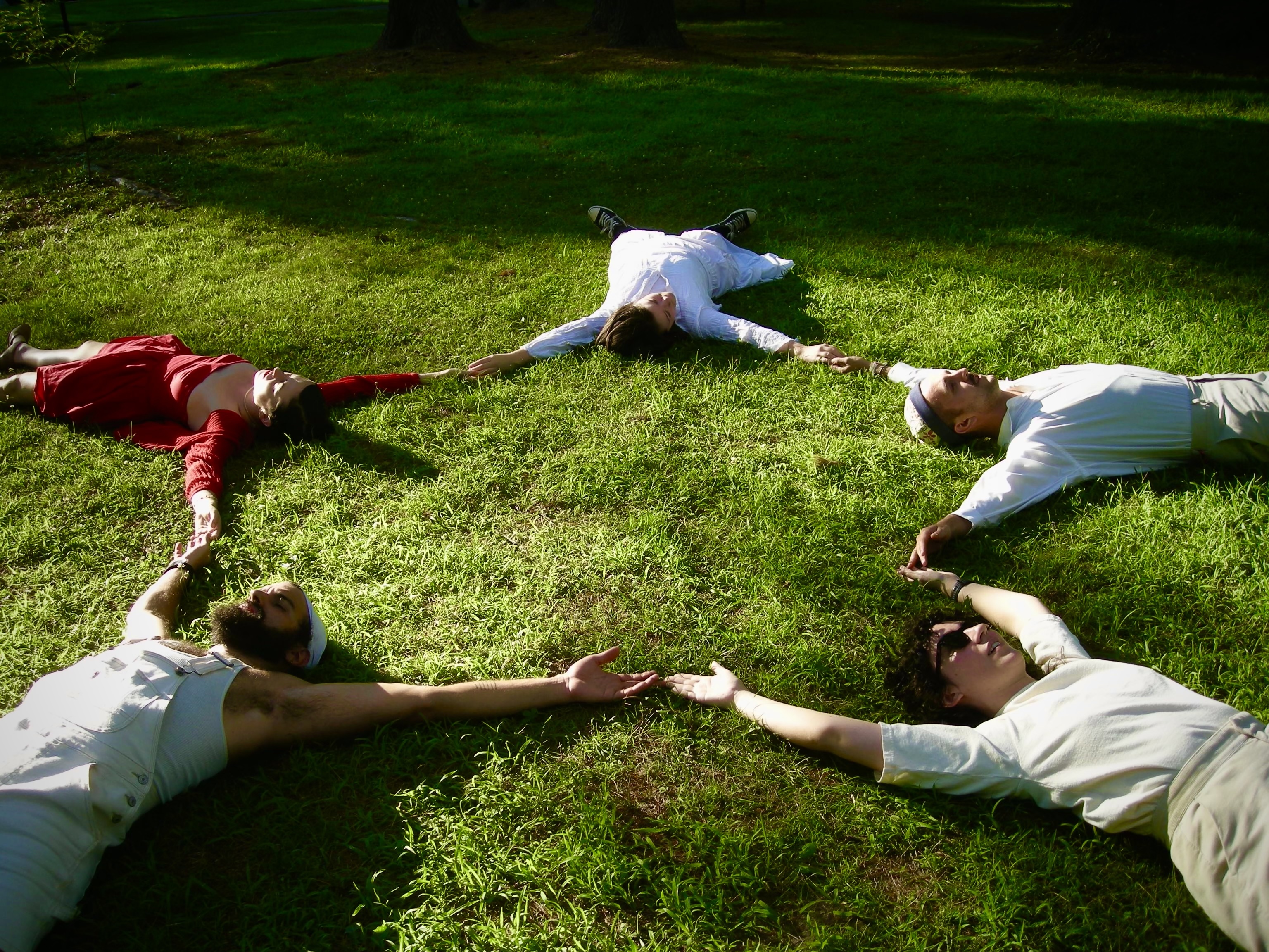 five people lying in green grass: Norma Jean Haynes Will Amend Camille Vogley Howes Madden Alea Nino Soberon and Eli Liguz. Norma Jean is dressed in red and everyone else is wearing white. Their arms make a star.